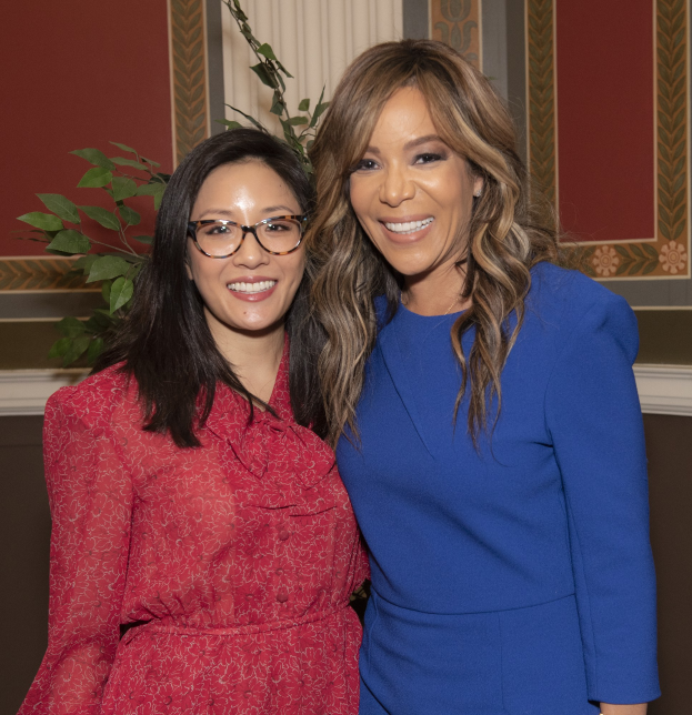 Zwei Frauen stehen nebeneinander und lächeln vor einer Wand; eine trägt ein blaues Kleid, die andere ein rotes Kleid mit Brille, mit einer Pflanze und einem Vorhang im Hintergrund bei der Oscar-Verleihung 2019 für Frauen im Film.