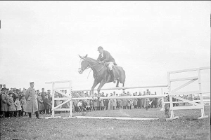 Schwarzes und weißes Foto eines Pferdes und Reiters, die über ein Hindernis springen, mit Zuschauern auf beiden Seiten.