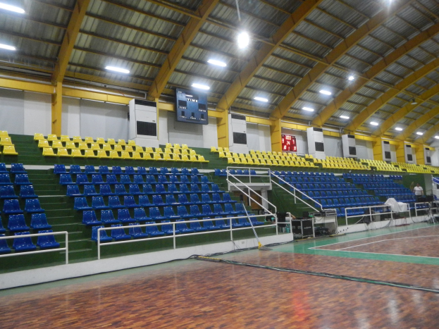 Großer Hallensportplatz mit Holzboden, blauen und gelben Sitzplätzen, Geländern, Treppe zum Sitzbereich, Wandbildschirmen, Deckenbeleuchtung und wenigen Anwesenden.