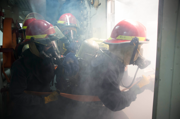 Eine Gruppe von Feuerwehrmännern in Helmen und Gasmasken steht in einem raucherfüllten Raum mit einer Tür auf der rechten Seite und verschiedenen Gegenständen im Hintergrund.