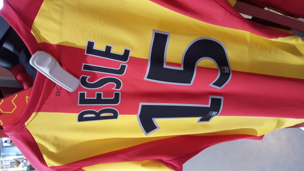 Ein roter und gelber Fußballtrikot mit Text und Nummern darauf, das an einer Stange in einem Geschäft hängt, zusammen mit anderen Gegenständen und dem Boden im Hintergrund.