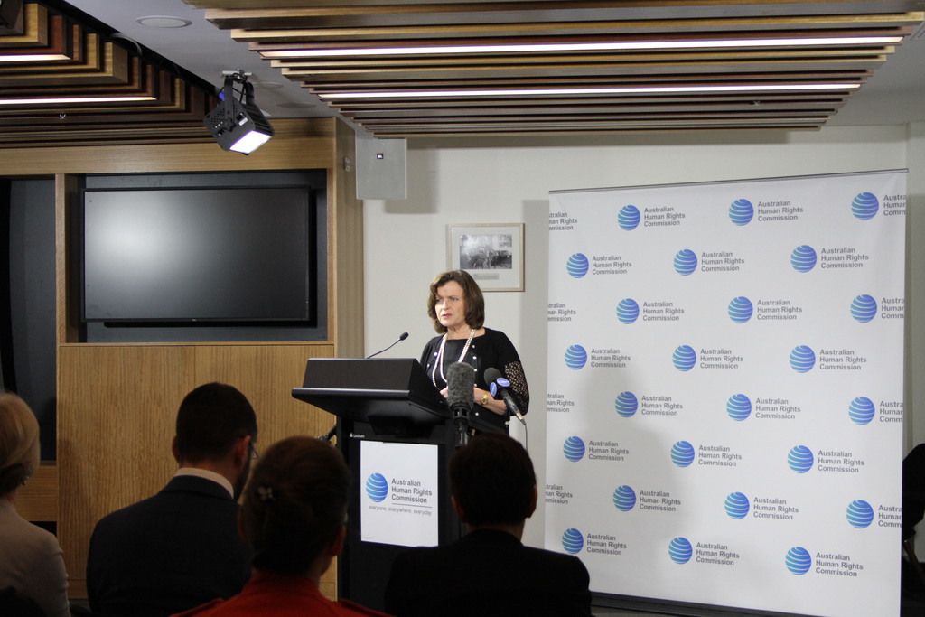 Eine Frau steht auf einer B├╝hne und spricht in ein Mikrofon, mit einem Publikum davor, einem Banner an der Wand dahinter, einem Fernseher an der Holzwand und Lichtern an der Decke.