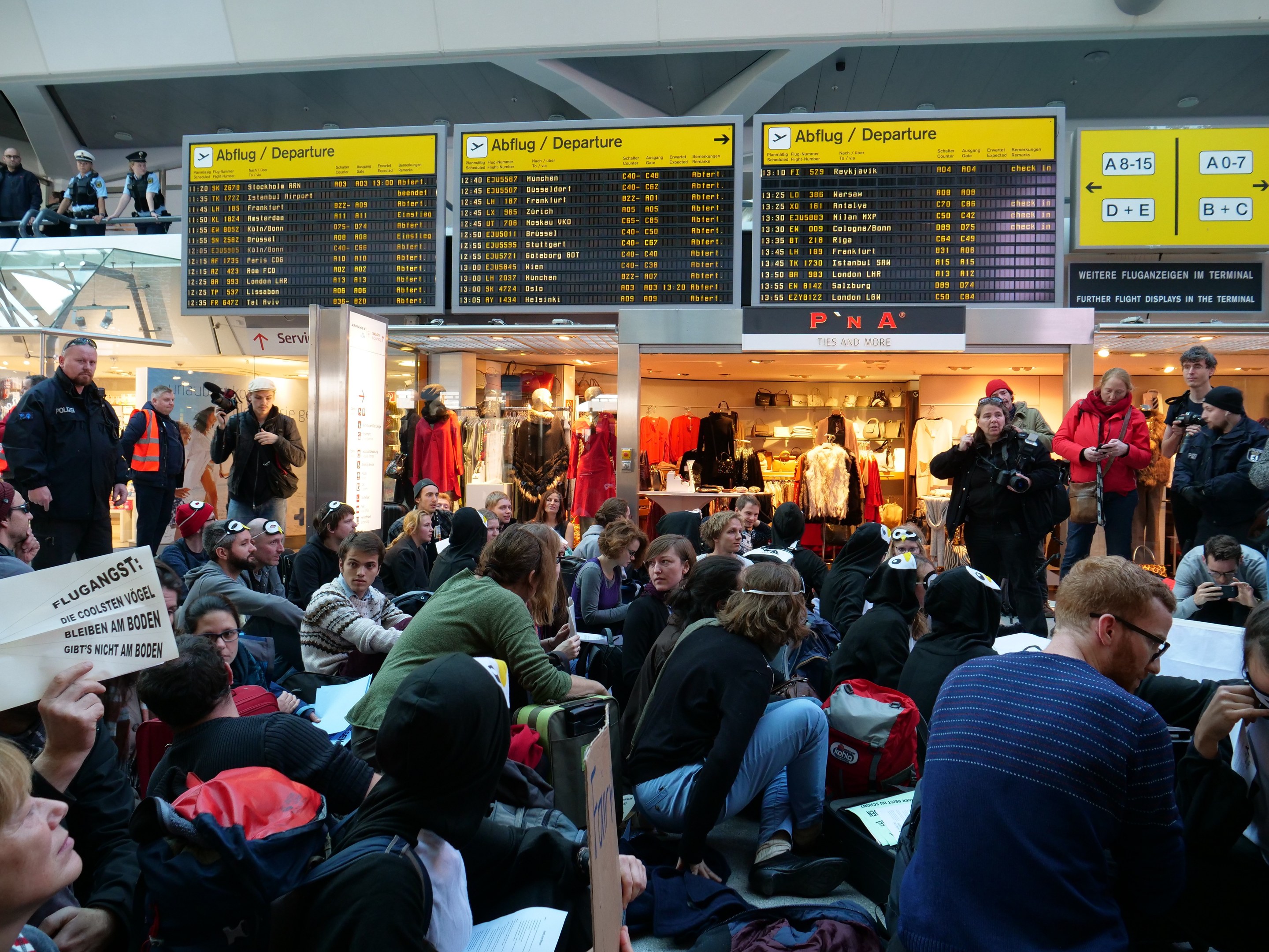 Eine große Gruppe von Menschen in einem Flughafen, einige sitzen mit Taschen und Papieren, andere stehen, mit Texttafeln, Schaufensterpuppen in Kleidern und Deckenbeleuchtung im Hintergrund, was auf eine Protestaktion hindeutet.
