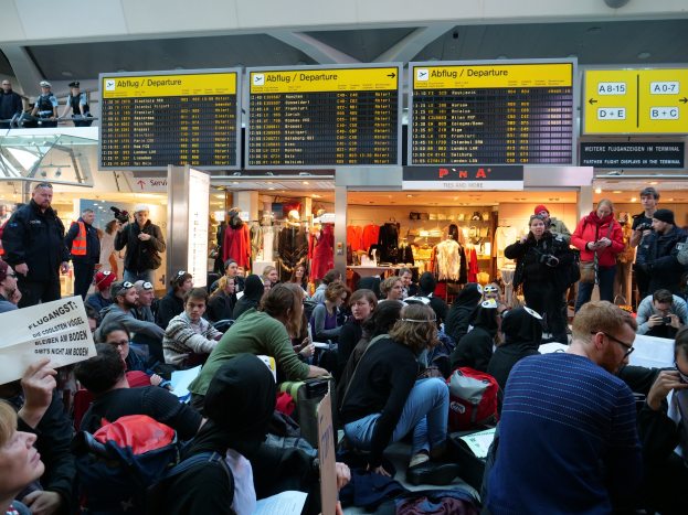 Eine große Gruppe von Menschen in einem Flughafen, einige sitzen mit Taschen und Papieren, andere stehen, mit Texttafeln, Schaufensterpuppen in Kleidern und Deckenbeleuchtung im Hintergrund, was auf eine Protestaktion hindeutet.