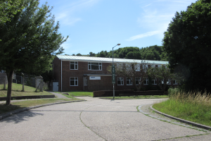 Ein Schulgebäude mit Fenstern, einem Straßenmast, einem Schild und einer Baumgruppe, umgeben von Gras und einem Zaun, unter einem bewölkten Himmel.