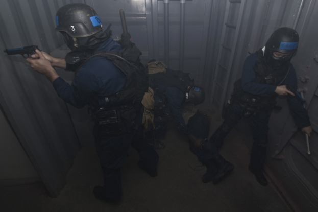 Eine Gruppe von Polizeibeamten in Einsatzausrüstung, einer hält eine Waffe, vor einem Metallcontainer und einer Leiter im Hintergrund.