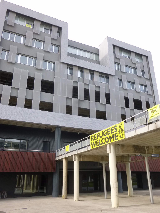 Großes Gebäude mit einem "Willkommen bei den Flüchtlingen"-Schild davor, umgeben von Säulen und einem Geländer, unter einem Himmel als Hintergrund.