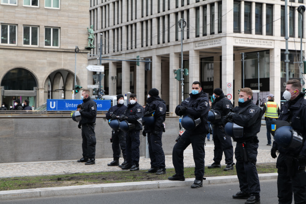 Eine Gruppe von Polizisten in schwarzen Uniformen und Masken steht vor einem Gebäude mit Glasfenstern und Säulen in Berlin, Deutschland, einige halten Helme, mit Laternenmästen, Verkehrsampeln, Schildern mit Text, einer Statue und anderen Menschen im Hintergrund auf einem grasbewachsenen Grund.