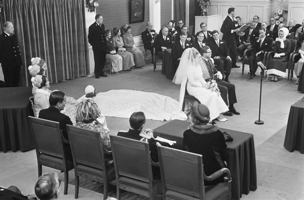 Schwarzes und weißes Foto einer Hochzeitszeremonie mit sitzenden und stehenden Gästen, einem Mann, der in ein Mikrofon spricht, einer Frau mit Blumen, einem Podium mit Mikrofon, Blumenschmuck, einem Vorhang, einem Rahmenfoto an der Wand und einer Tür im Hintergrund.