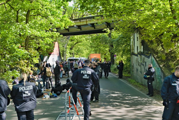 Polizeibeamte hinter einer Barriere mit Menschen, die auf der Straße sitzen und stehen, Bäumen und einer Brücke im Hintergrund und einer Wand mit Schrift auf der rechten Seite.