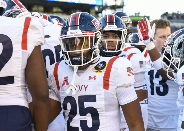 Gruppe von Männern in Football-Uniformen auf einem Feld mit Helmen und Handschuhen, Hintergrund zeigt eine unscharfe Menge, ein Scoreboard mit "Auburn vs Virginia Tech - 11/28/15 College Football Pick, Odds, and Prediction" und Himmel.