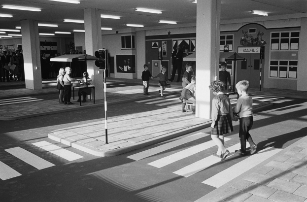 Schwarß-weißes Foto von Kindern, die eine Straße an einer Ampeläberquerung mit nahen Passanten überqueren, mit einem Verkehrssignalmast im Vordergrund und strukturellen Pfeilern mit Deckenleuchten im Hintergrund.