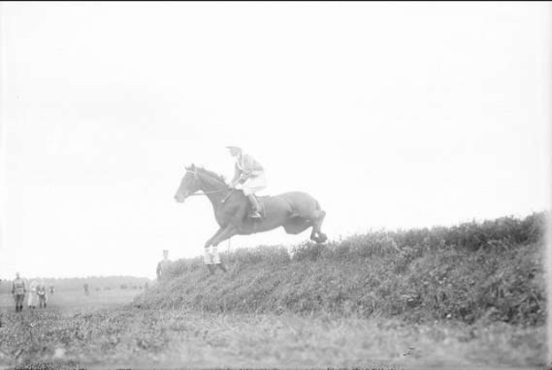 Ein Schwarz-Weiß-Foto eines Pferdes und Reiters, die über einen grasbewachsenen Hügel springen, mit dem Pferd im Galopp und dem Reiter in einem Helm und einem langen Mantel, während Zuschauer im Hintergrund beobachten.