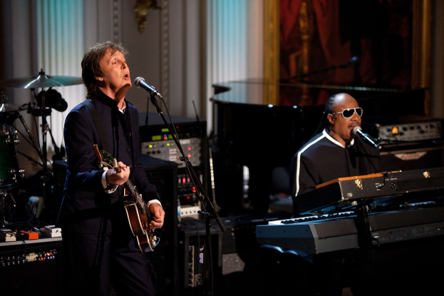 Paul McCartney spielt Gitarre und singt in ein Mikrofon, während Stevie Wonder eine Brille trägt und am Piano auf der Bühne der Grammys spielt, mit Instrumenten und einem gerahmten Bild im Hintergrund.