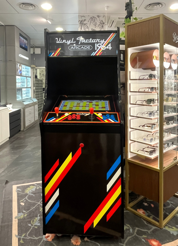 Ein Geschäft mit einem zentralen Retro-Arcade-Automaten, einem rechten Gestell mit Brillen und Hintergrund-Elementen wie Schränken, einem Fernseher und anderen Gegenständen, mit Deckenleuchten, die eine Retro-Atmosphäre schaffen.