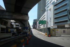 Eine Stadtstraße mit Verkehrskegeln am Straßenrand, Fahrzeugen auf der Straße, einer Brücke auf der linken Seite, Gebäuden mit Namensschildern auf beiden Seiten, Laternenmasten und einem Himmel im Hintergrund.