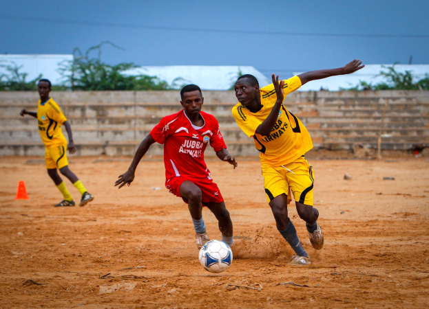 Eine Gruppe von Männern in bunten Trikots, die auf einem Feld mit einer Wand, Bäumen und einem klaren blauen Himmel Fußball spielen.