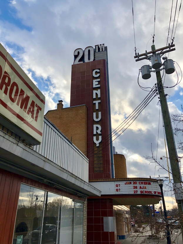 Außensicht des 20. Jahrhundert-Theaters in Detroit, Michigan, mit Glaswänden, Namensschildern, umgeben von Bäumen und Strommästen mit Drähten unter einem bewölkten Himmel.