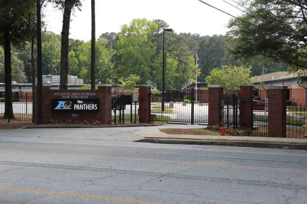 Eingang einer Schule mit einem Schild, umgeben von B├Ąumen, Stra├čenm├Âbeln, Fahrzeugen und Geb├Ąuden unter einem klaren blauen Himmel.