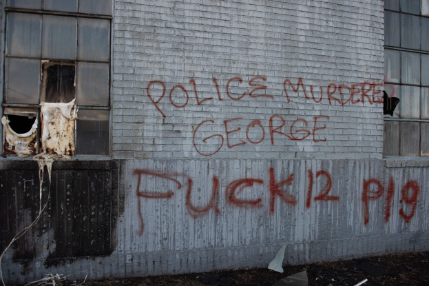 Ein Gebäude mit der Graffiti-Beschriftung "Polizei ermordet George Fuck 12 P19" an der Seite, das Fenster und verschiedene Gegenstände vorne zeigt.