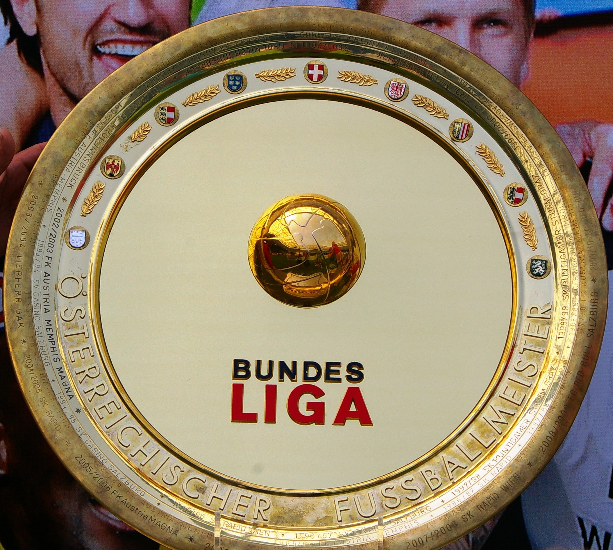 Ein Mann hält einen goldenen Schild mit "Bundesliga" darauf, im Hintergrund eine Fahne mit drei Personen.