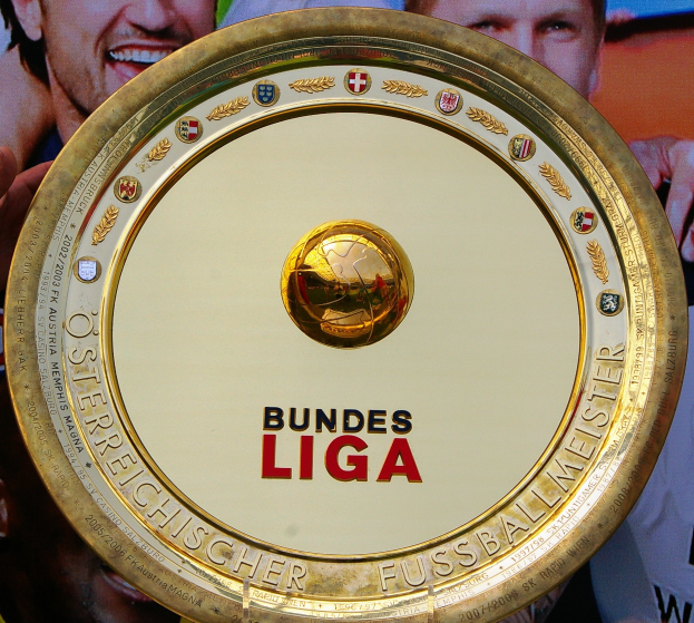 Ein Mann hält einen goldenen Schild mit "Bundesliga" darauf, im Hintergrund eine Fahne mit drei Personen.