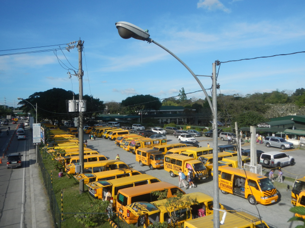 Eine große Anzahl gelber Schulbusse entlang einer Straße mit Fußgängern auf dem Gehweg, Strommasten, Bäumen, Gebäuden und einem bewölkten Himmel im Hintergrund.