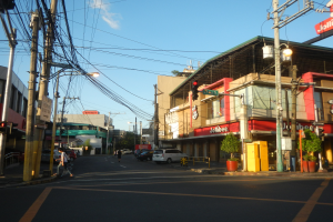 Eine belebte Stadtstraße mit Fußgängern, Fahrzeugen und verschiedenen städtischen Elementen wie Ampeln, Gebäuden und Pflanzen unter einem bewölkten Himmel.