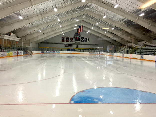 Große Indoor-Eisbahn mit Deckenbeleuchtung, Werbetafeln, einer Wand, Treppen mit Geländern und einigen Menschen, wahrscheinlich eine Hockeyanlage.
