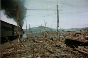 Ein Zug fährt durch ein beschädigtes Gebiet mit Rauch, der aus ihm aufsteigt, umgeben von Strommasten und -leitungen, mit Hügeln und einem klaren Himmel im Hintergrund und verstreutem Schutt auf dem Boden.