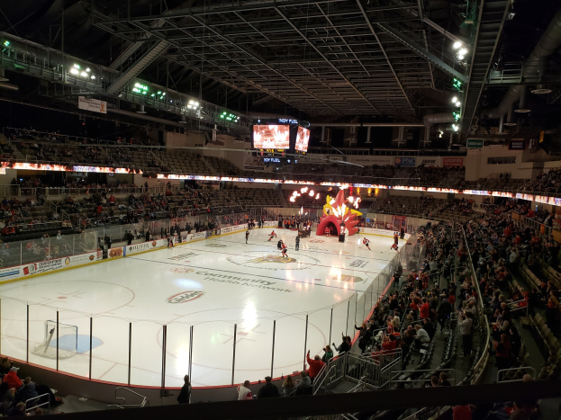 Eishockeyspiel in einer großen Arena mit Sitz- und Stehplätzen in der Nähe der Eisfläche, umgeben von Bannern, Metallrohrkonstruktionen und zwei Überwachungsschirmen, identifiziert als Columbus Blue Jackets Arena.