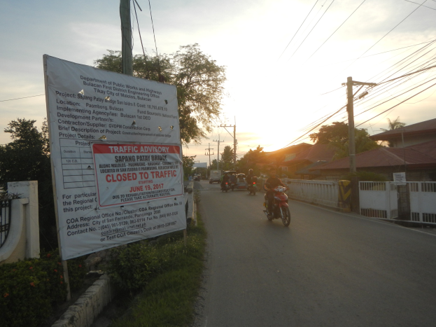 Eine Gruppe von Motorradfahrern fährt auf einer Straße mit einem "Verkehrshinweis Geschlossen für den Verkehr"-Schild, gesäumt von einer blumenbedeckten Wand und Bäumen, mit Häusern und einem klaren Himmel im Hintergrund.