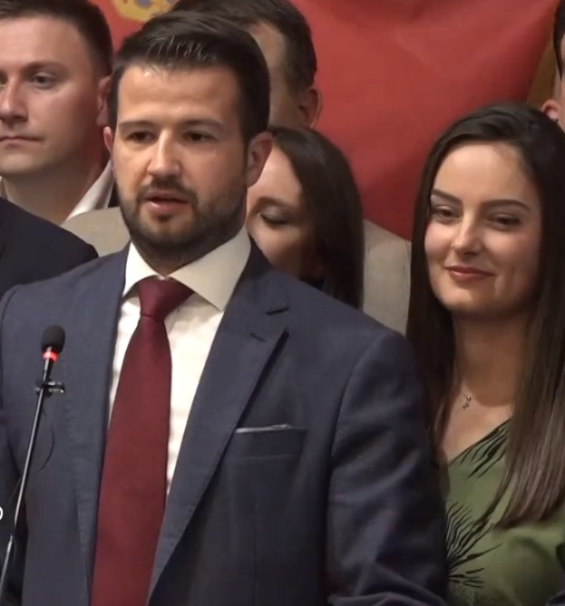 Mann in Anzug und Krawatte, der vor einem Publikum an einem Rednerpult spricht und ein Mikrofon hält, mit einem Banner im Hintergrund.