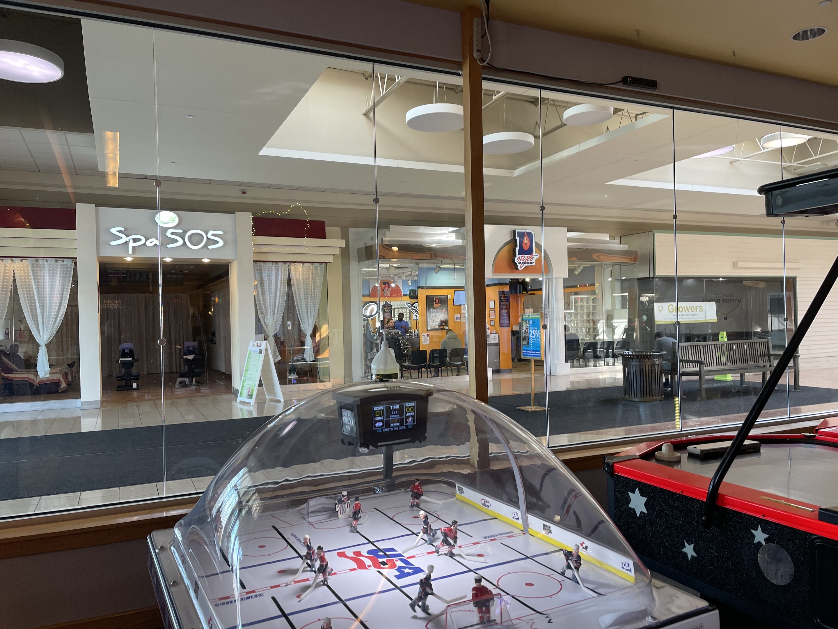 Eine Eishalle in einem Einkaufszentrum, umgeben von Glaswänden mit Menschen, Möbeln und Beschriftungen draußen sichtbar und einem Air-Hockey-Tisch auf der rechten Seite.