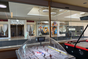 Eine Eishalle in einem Einkaufszentrum, umgeben von Glaswänden mit Menschen, Möbeln und Beschriftungen draußen sichtbar und einem Air-Hockey-Tisch auf der rechten Seite.