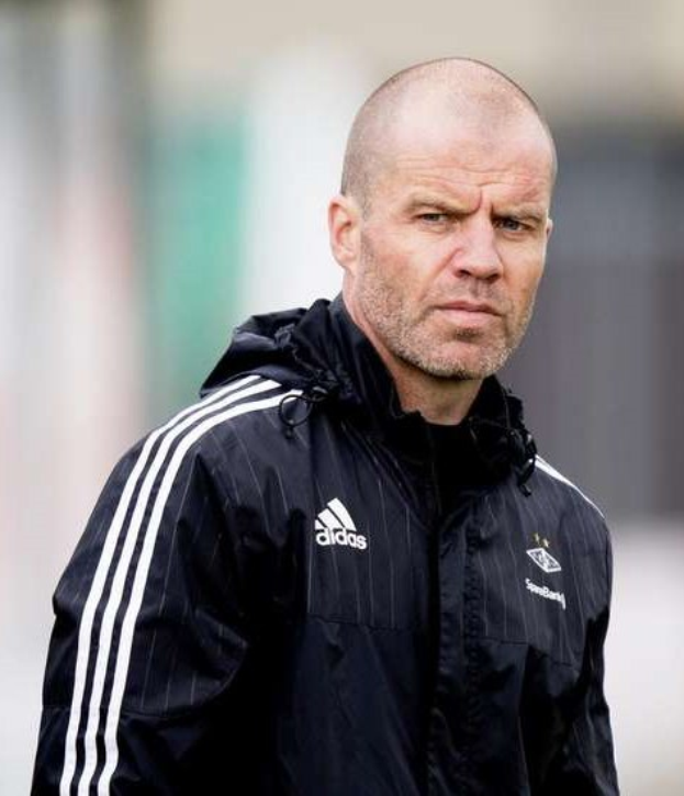 Ein kahlköpfiger Mann in einer schwarzen Jacke steht auf einem Fußballfeld, trägt eine marineblaue Jacke mit weißen Streifen und einem weißen Kragen, die Hände sind verschränkt und der Gesichtsausdruck ernst.