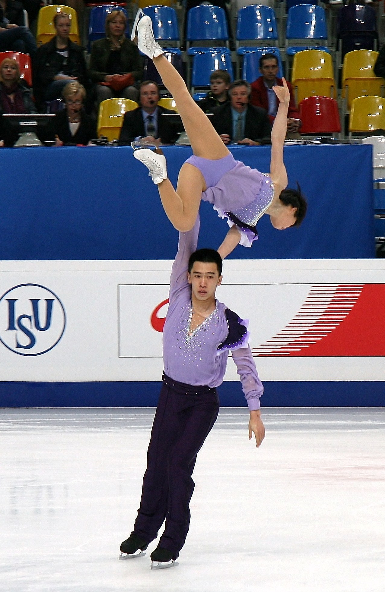 Zwei Eiskunstläuferinnen, eine in einem violetten Kleid und eine in einem weißen Kleid, performen auf einer Eisbahn mit Zuschauern und einem blauen und weißen Schild im Hintergrund.