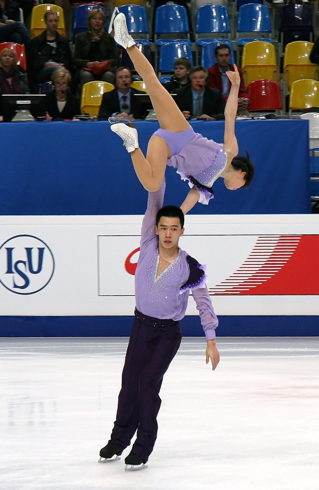 Zwei Eiskunstläuferinnen, eine in einem violetten Kleid und eine in einem weißen Kleid, performen auf einer Eisbahn mit Zuschauern und einem blauen und weißen Schild im Hintergrund.