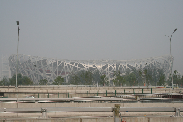 Olympiastadion in Peking, China, mit Brücke, Geländern, Pfählen, Laternen, Bäumen und einem Gebäude im Hintergrund vor einem klaren blauen Himmel.