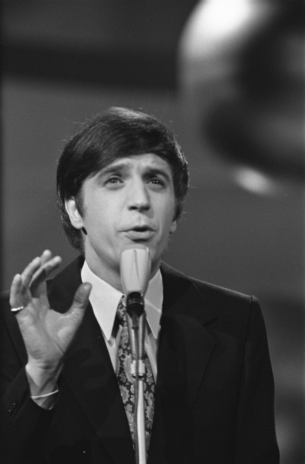 Ein Mann in einem schwarzen Anzug, weißem Hemd und gemustertem Schlips spricht in ein Mikrofon, mit ernster Miene und zurückgekämmtem Haar, vor einem unscharfen Hintergrund.