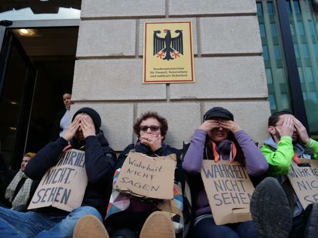 Eine Gruppe von Menschen sitzt vor einem Gebäude und hält Schilder, während einige ihr Gesicht verdecken.