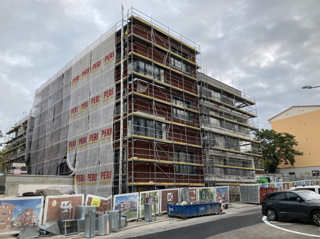 Großes Gebäude im Bau mit Gerüst, umgeben von Bäumen, Straßeninfrastruktur und Fahrzeugen, vor einem bewölkten Himmel.