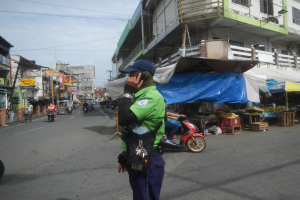 Ein Polizist in grüner und blauer Uniform mit blauer Mütze steht auf einer Straße, spricht in ein Handy, mit Fahrzeugen und Marktbuden im Hintergrund und einem Gebäude mit Fenstern im bewölkten Hintergrund.