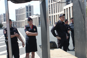 Eine Gruppe von Polizisten in Uniform auf einer Straße mit Masten und Kabeln im Vordergrund, Gebäuden, Bäumen und einem klaren blauen Himmel im Hintergrund.
