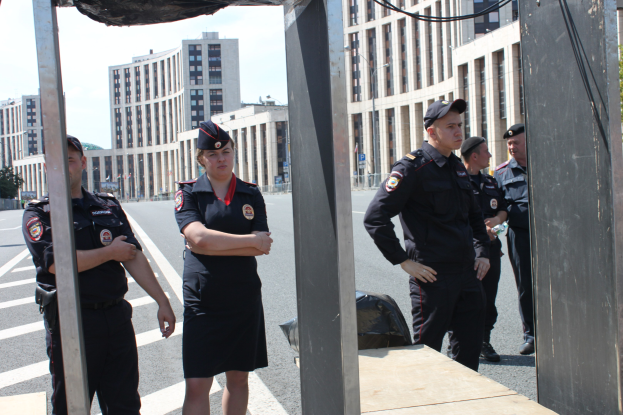 Eine Gruppe von Polizisten in Uniform auf einer Straße mit Masten und Kabeln im Vordergrund, Gebäuden, Bäumen und einem klaren blauen Himmel im Hintergrund.