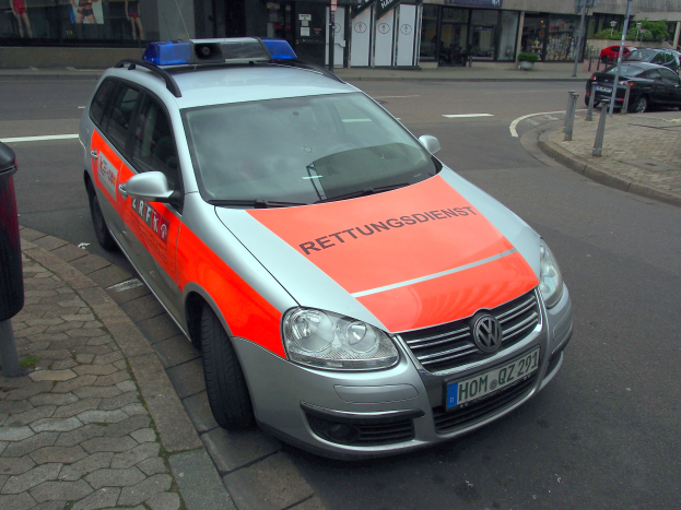 Polizeifahrzeug am Straßenrand geparkt mit einem Müllcontainer auf dem Gehweg links, Gebäude und Bäume im Hintergrund.