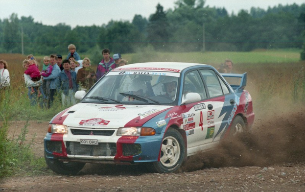 Ein Rallyeauto mit Text und Zahlen fährt eine Schotterstraße entlang, umgeben von einer Menschenmenge, einige halten Kameras, mit Pflanzen, Gras, Polen, Drähten, Bäumen und einem klaren blauen Himmel im Hintergrund.