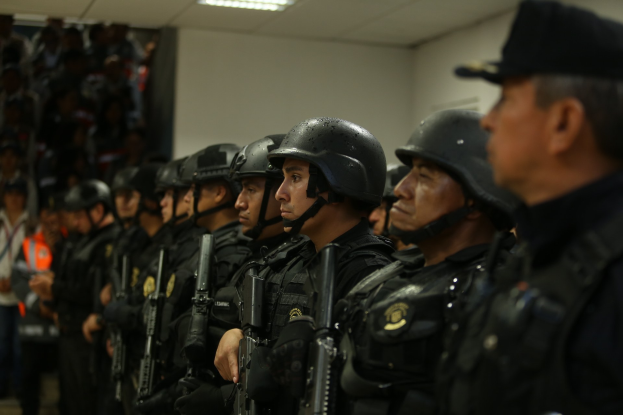 Gruppe von Polizisten in Uniform, die nebeneinander stehen und alle Helme tragen und Waffen halten, mit einer Wand und Deckenbeleuchtung im Hintergrund.