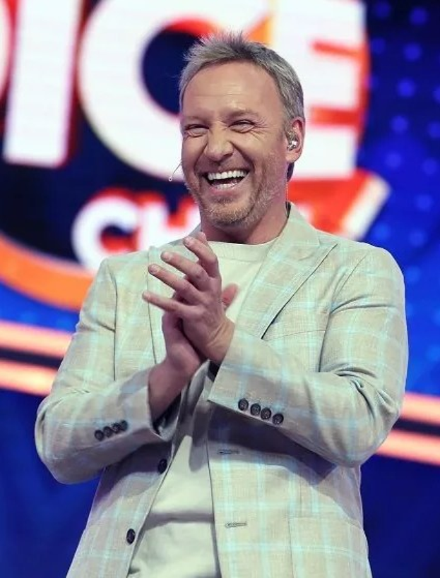 Ein Mann in einem Anzug, der auf einer Bühne klatscht, mit 'The Voice'-Logo im Hintergrund.