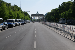 Eine Reihe von Polizeiwagen auf einer Straße vor dem Brandenburger Tor in Berlin, Deutschland geparkt, mit Menschen auf Fahrrädern und Stehenden, Barrieren, Bäumen und einem Bogen mit Statuen im Hintergrund.